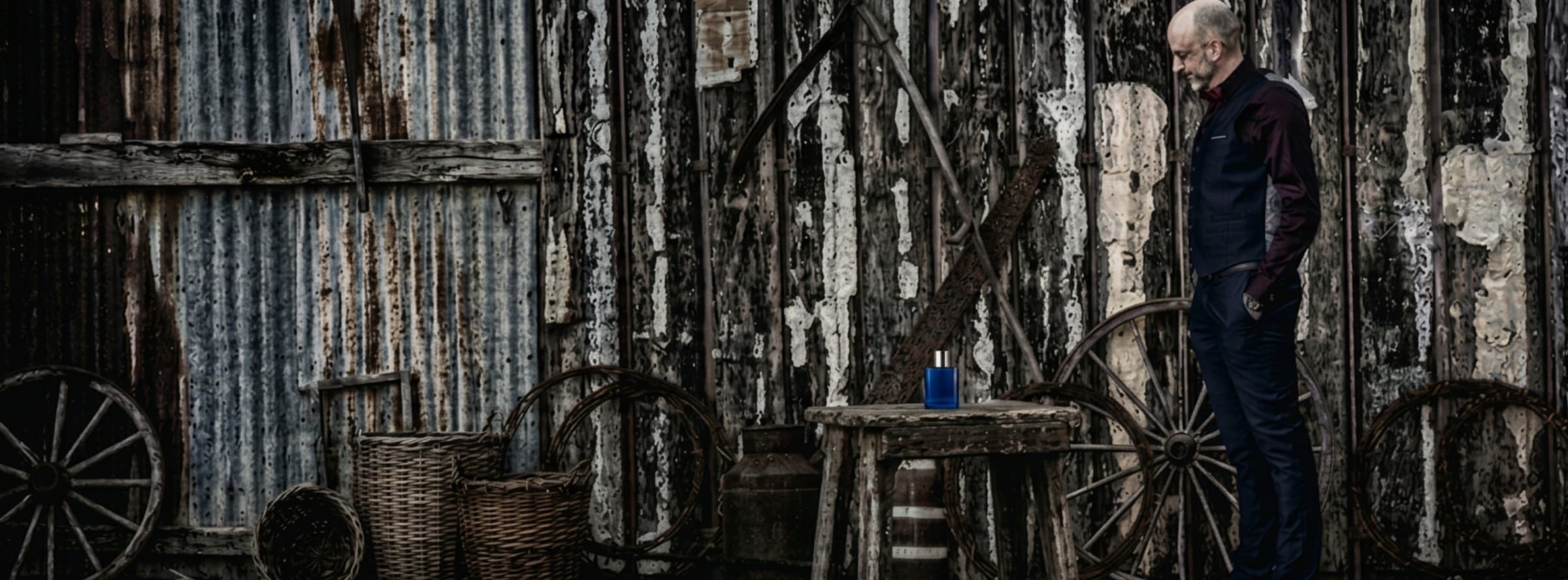 A man beside a weathered corrugated-iron shed, a blue bottle standing on an old wooden stool